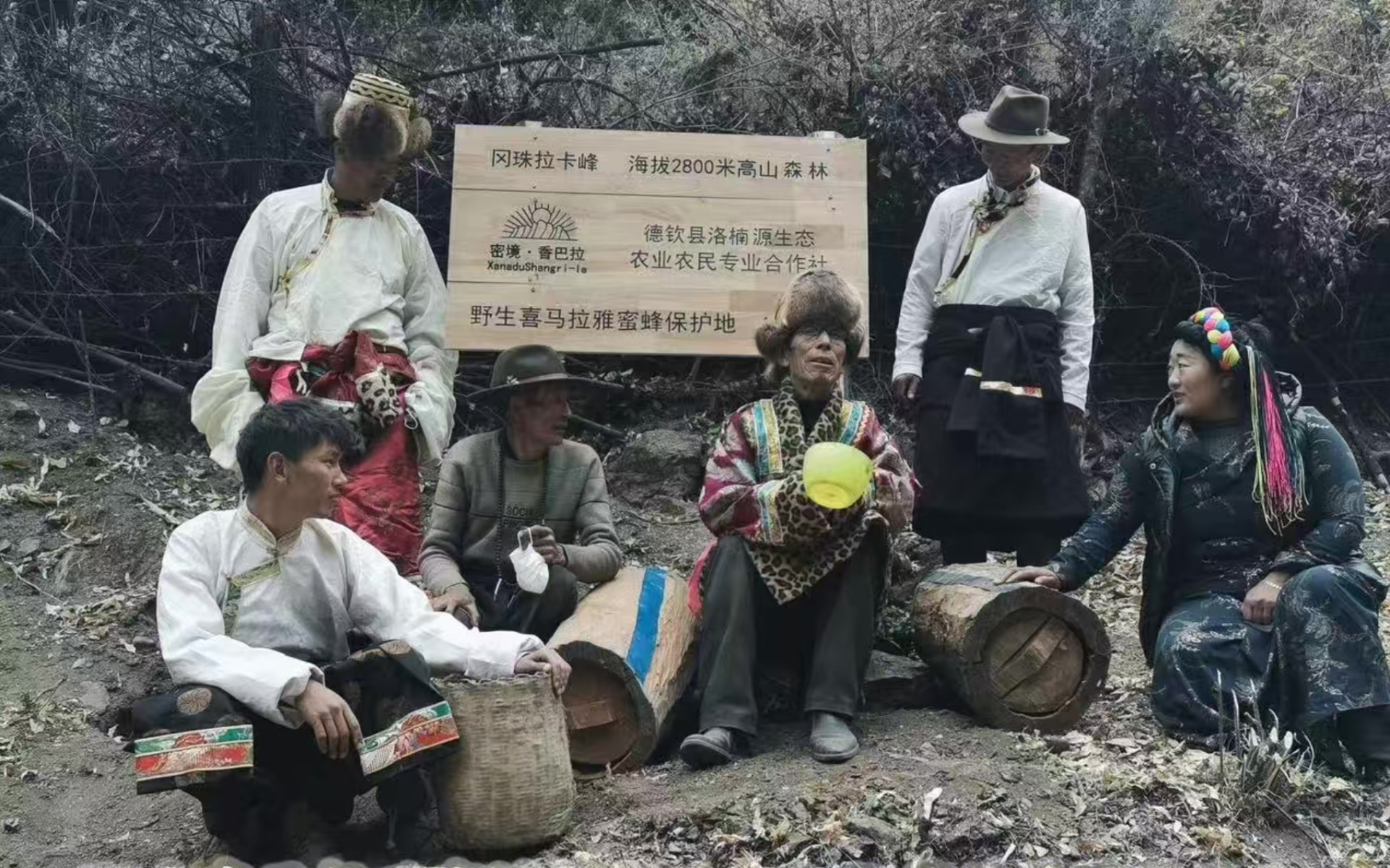 Seven Tibetan beekeeping inheritors gathered with traditional hollow-log hives in front of the Wild Himalayan Honeybee Conservation Area signboard